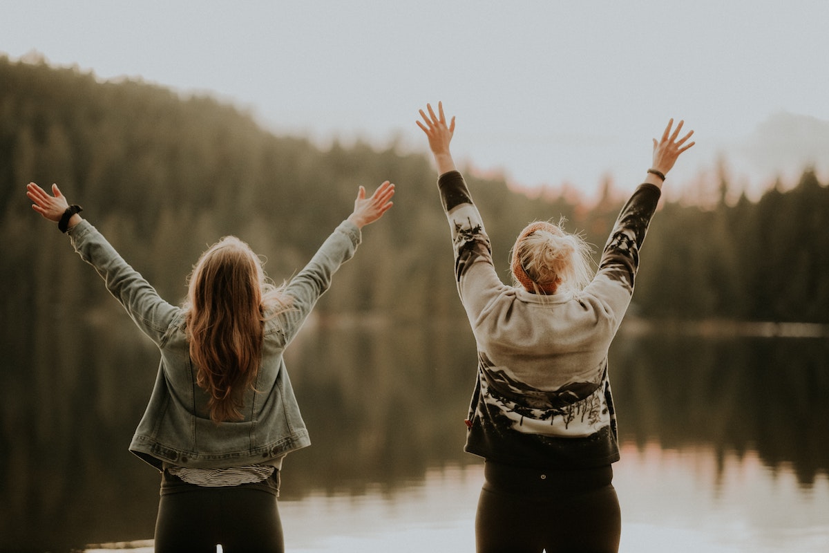 Two women facing a body of water with their arms happily outstretched.