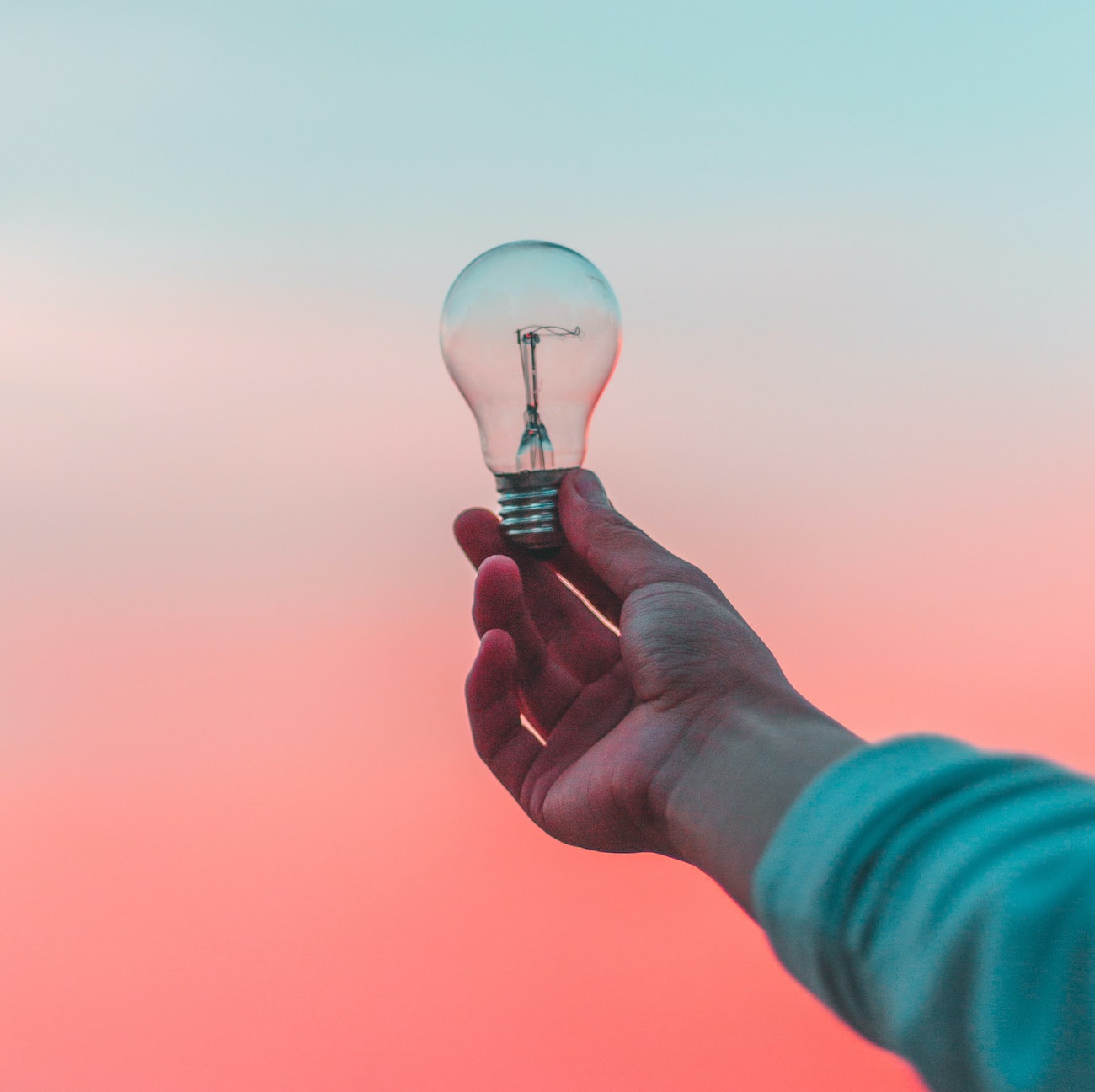 Close-up of an outstretched hand holding a lightbulb in front of a pinkish, orange sunset.