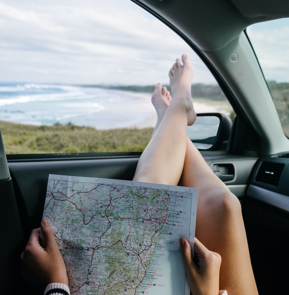 A person laying in a car, holding a map, and sticking their feet out the window.